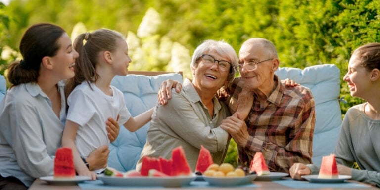 Loving Family Embracing Elderly Grandparents at Outdoor Table Near Memory Care Facility